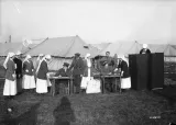 Photographie en noir et blanc d’infirmières et d’hommes en uniforme de soldats devant une série de tentes. À droite, une infirmière se tient derrière un écran de vote. Seules sa tête et ses épaules sont visibles.