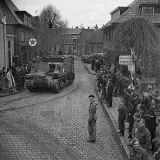 Une foule assemblée sur des trottoirs et regardant des véhicules blindés rouler sur les rues pavées d’un village.