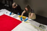 Deux femmes portant des gants et des tabliers de protection mesurent la longueur d’un grand drapeau du Canada, posé à plat sur une table.