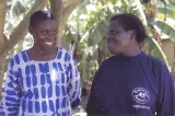 Deux femmes, debout devant des arbres tropicaux, se regardent et sourient. La femme de droite porte un chandail qui porte le logo de la Concerned Parents Association.