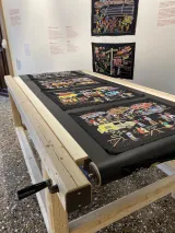 Une série de panneaux de broderie colorés sur du tissu noir, exposés sur une table de convoyeur en bois à manivelle et sur le mur d’un musée.