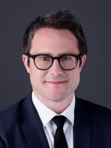 Headshot of a smiling man with short, dark-brown hair. He is wearing glasses and a suit.