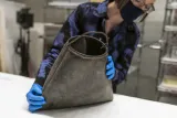 Une femme tient un sac en caoutchouc noir éraflé avec une poignée métallique courbée de forme grossière sur le dessus.