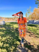 Une jeune femme est représentée portant une robe orange à clochettes. Elle est dehors dans un parc. On peut voir une statue d'avion derrière elle.