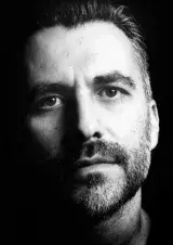 Homme avec une moustache et une courte barbe. Photographié en noir et blanc.