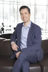 Un homme caucasien en costume d'affaires a les cheveux bruns et les yeux bleus. Il est assis les jambes croisées.