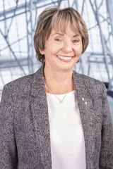 Une femme blanche aux cheveux châtain clair porte un blazer gris à motifs et une chemise blanche. Elle sourit face à la caméra et se tient debout de manière détendue.