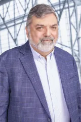 Un homme avec cheveux gris, porte un blazer bleu-mauve avec une chemise blanche.