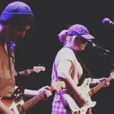 Deux guitaristes, l’un portant une tuque et l’autre une casquette de baseball, jouent sur scène sous une lumière violette. Un microphone et une autre guitare sont visibles en bordure.