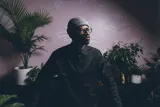 Alpha Toshineza, un homme noir portant une tuque, des lunettes rondes et une expression intense, se tient dans une pièce remplie de plantes tropicales.