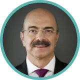 Un homme à la moustache sombre et aux petites lunettes sans monture porte un complet sombre et une cravate violette.