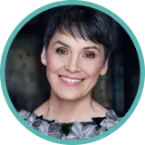 Une femme aux cheveux sombres et courts, vêtue d’un chemisier à collier de fleurs, sourit.