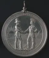 Médaille ronde de couleur argentée portant l’inscription « INDIAN TREATY No. » en haut et « 187 » en bas. La partie centrale montre un lever de soleil lointain et des tipis derrière deux personnages qui se serrent la main. À gauche, un homme vêtu d’une tenue militaire coloniale et à droite, un homme torse nu portant un pantalon à franges et une coiffe de plumes.