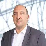 Un homme blanc porte un blazer gris foncé et une chemise blanche. Il fait face à la caméra et affiche un léger sourire.