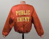 Le dos d’une blouson style aviateur de couleur rouge-orange foncé, ornée de l’inscription « Public Enemy » en grandes lettres majuscules jeunes.