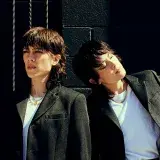 Deux personnes portant des chemises blanches et des blazers noirs posent contre un mur en parpaings peint en noir.