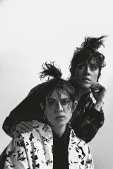 Deux personnes aux cheveux longs attachés en queue de cheval. L'une se tient derrière l'autre et s'appuie sur ses épaules. Elle porte une chemise à motifs noirs et blancs sous une veste en cuir noir. Devant, l'autre porte une chemise noire unie sous une veste à motifs noirs et blancs.