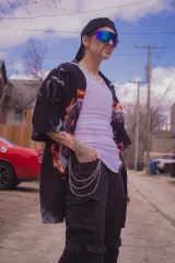 Une artiste de drag porte une casquette, une chemise et un pantalon noirs avec des chaînes, ainsi que des lunettes bleues et un débardeur blanc.