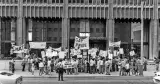 Un groupe de personnes manifeste sur le trottoir devant un immeuble de bureaux. Elles tiennent des pancartes et des banderoles de protestation et sont disposées sur trois ou quatre rangs. Un policier les observe.