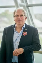 Un homme se tient face à l’objectif avec une expression sérieuse. Il porte une veste de costume sombre et une chemise boutonnée bleu clair. Il arbore une fleur multicolore de style métis sur son revers.