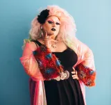 Une artiste de drag aux cheveux roses volumineux et au maquillage noir sur les yeux et les lèvres pose une main sur la hanche. Elle porte une veste rose en voile avec des poignets en fourrure colorée par-dessus une robe noire.