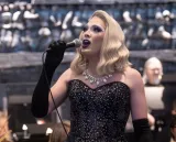 Une élégante artiste de drag blonde, vêtue d’une robe noire à paillettes, d’un collier de diamants et de longs gants de soirée noirs, chante dans un microphone.