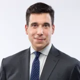 Un homme blanc aux cheveux foncés sourit légèrement à la caméra. Il porte un costume sombre avec une chemise blanc cassé et une cravate bleu foncé à petits pois blancs.