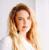 Une femme blanche aux longs cheveux blonds et portant un anneau dans le nez regarde la caméra. Elle porte une chemise blanche.