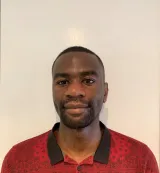 Une personne noire aux cheveux noirs très courts regarde la caméra avec un léger sourire. Elle porte une chemise rouge avec un motif imprimé représentant un dessin au trait noir et un col noir.