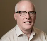 Un homme d’âge moyen aux cheveux gris courts et portant des lunettes, vêtu d’une chemise blanche et souriant chaleureusement vers la caméra.