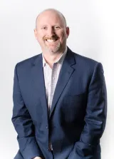 Un homme blanc aux cheveux courts et à la barbichette sourit à la caméra. Il porte un costume bleu foncé et une chemise à carreaux blancs.