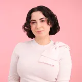 Une femme blanche aux cheveux bruns sourit à la caméra. Elle porte un gilet rose pâle avec un nœud rose sur l’épaule gauche.