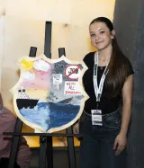 Une jeune femme se tient à côté d’un bouclier peint représentant d’un côté un ciel ensoleillé et un bateau flottant sur l’eau, et de l’autre un ciel sombre et un bateau en train de couler.