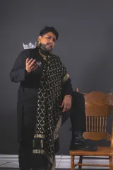Un drag king barbu, vêtu d'une tenue noire et d'une longue écharpe à motifs noirs et dorés, lève la main droite, une couronne sertie de pierres précieuses posée sur son épaule, et prend la pose avec assurance devant un mur gris, à côté d'une chaise en bois.