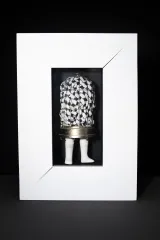 Une sculpture en techniques mixtes composée d’une cage à oiseaux métallique au sommet arrondi, avec des jambes de poupée blanches fixées à la base. La cage renferme un tissu blanc orné de motifs noirs. L’ensemble est présenté dans un cadre blanc.