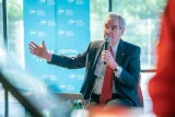 Un homme en complet et cravate rouge parle dans un micro et fait des gestes, assis devant une bannière de la Central European University.