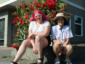 Tiana Garcia et Megumi Kimata sont assises, souriantes, devant un rosier rouge. Tiana Tiana a de longs cheveux fuchsia et des lunettes de soleil roses. Megumi a des cheveux foncés et porte des lunettes rondes et un chapeau.