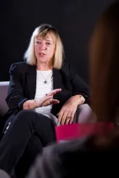 Une femme assise dans un fauteuil. Son coude gauche est posé sur un accoudoir et sa main gauche est étendue devant elle alors qu’elle parle.