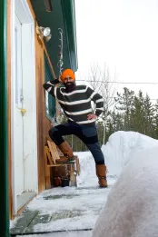 Un homme sikh canadien, souriant, portant un pull rayé noir et blanc, un pantalon bleu et marron, des bottes et un turban orange, dans la neige est appuyé sur le côté d'une cabane.