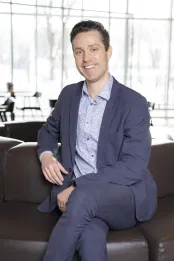Un homme caucasien en costume d'affaires a les cheveux bruns et les yeux bleus. Il est assis les jambes croisées.