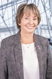Une femme blanche aux cheveux châtain clair porte un blazer gris à motifs et une chemise blanche. Elle sourit face à la caméra et se tient debout de manière détendue.
