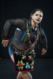 Melody McKiver, Anishinaabe, porte un chemisier noir transparent sur un débardeur arc-en-ciel, du rouge à lèvres noir et une expression intense. Iel se penche vers l’avant, ses cheveux noirs en deux tresses, avec un carré de tissu brodé porté en bandoulière sur une lanière perlée.