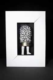 Une sculpture en techniques mixtes composée d’une cage à oiseaux métallique au sommet arrondi, avec des jambes de poupée blanches fixées à la base. La cage renferme un tissu blanc orné de motifs noirs. L’ensemble est présenté dans un cadre blanc.