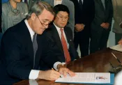 Brian Mulroney assis à une table, signant un document. M. Art Miki est assis à ses côtés et l’observe.