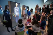 A woman stands in a museum gallery beside a glowing, full-sized image of Nelson Mandela. She is speaking to a crowd of children and adults who are gathered around her.
