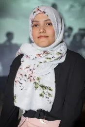 Un portrait d’une jeune femme aux yeux foncés qui porte une chemise noire à manches longues. Ses cheveux sont couverts d’un foulard principalement blanc avec un motif floral vert et rose.