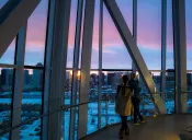 Deux personnes regardent par une grande fenêtre un coucher de soleil sur un paysage urbain.