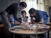 Une femme et un homme se penchent au-dessus d'une table où un jeune enfant joue avec une pile de photographies.
