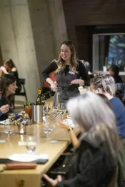 Women drink wine around a table in a restaurant. They are being served by a smiling woman in an apron.