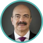 Un homme à la moustache sombre et aux petites lunettes sans monture porte un complet sombre et une cravate violette.